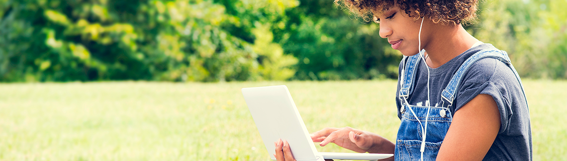 Woman using laptop computer, with headphones in her ears, sitting outside in a grassy area.